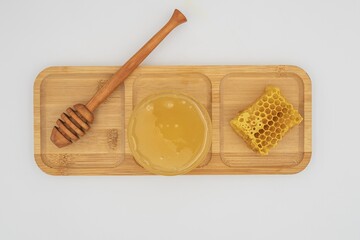 Glass bowl filled with honey, honeycomb piece and wooden dipper on bamboo tray. Clean white background, natural sweet food and organic ingredient concept.