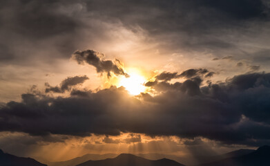 Golden sunlight through dark clouds over mountains
- sunset sky with golden sun rays breaking through dark storm clouds, creating a powerful contrast of light and shadow over distant mountains.