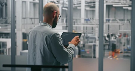 Bearded Male Engineer Analyzing Production Data Graphs on Tablet. Man Standing in Middle of Modern, Automated Manufacturing Facility. Concept of Mobile Data Analysis and Smart Factory Operations. - Powered by Adobe