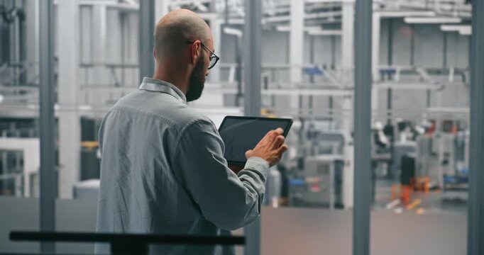 Bearded Male Engineer Analyzing Production Data Graphs on Tablet. Man Standing in Middle of Modern, Automated Manufacturing Facility. Concept of Mobile Data Analysis and Smart Factory Operations.