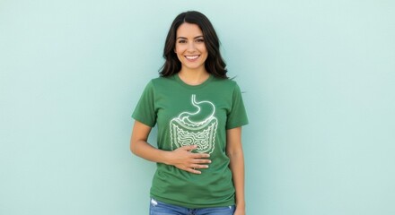 A smiling brunette woman wearing a green t-shirt with a digestive system graphic stands against a light blue wall in natural lighting. She is holding her abdomen area.