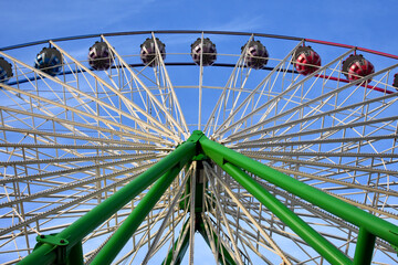 Low-angle perspective of a giant ferris wheel against a clear blue sky. The image highlights the intricate white steel spoke structure and colorful gondolas radiating from a vibrant green hub.