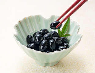 Picking Up a Glossy Sweetened Black Soybean (Kuromame) with Chopsticks from an Elegant Shell-Shaped Bowl, Traditional Japanese New Year Dish with Copy Space