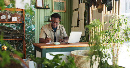 Laptop, writing and plant with black man in store for delivery checklist, account report and botany review. Order confirmation, ecommerce and florist entrepreneur with person in small business