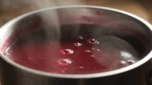 Close up of a stainless steel saucepan filled with simmering, steaming dark red liquid, possibly a fruit-based compote or reduction