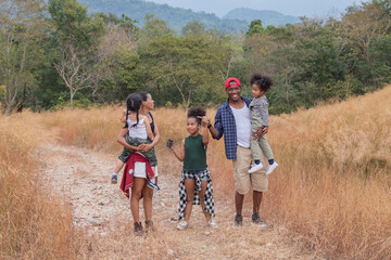 Obraz premium Happy African American family with adorable children weekend vacation hiking outdoors in countryside, father and mother carry children walking trail in forest park with fun, bonding relationship