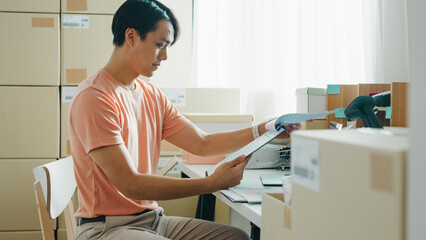 Candid of young attractive Asia male entrepreneur E-commerce small business check sale purchase order from customer in document paper and inventory product ready shipping at small online retail shop.