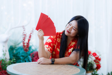 Portrait of happy asian woman relaxing wearing red cheongsam dress holding red envelopes at home in the living room indoor decoration for Chinese new year festival celebrate culture of china holiday