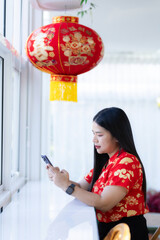 Portrait of asian woman wearing red cheongsam dress holding red envelopes and smartphone at home in the living room indoor decoration for Chinese new year festival celebrate culture of china holiday