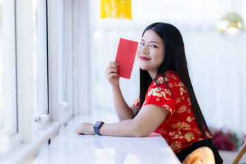 Portrait of happy asian woman relaxing wearing red cheongsam dress holding red envelopes at home in the living room indoor decoration for Chinese new year festival celebrate culture of china holiday