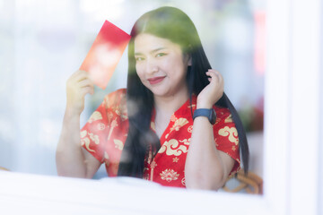 Portrait of happy asian woman relaxing wearing red cheongsam dress holding red envelopes at home in the living room indoor decoration for Chinese new year festival celebrate culture of china holiday