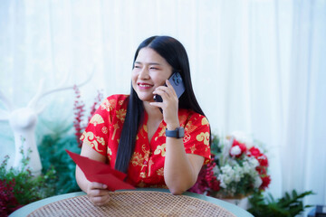 Portrait of asian woman wearing red cheongsam dress holding red envelopes and smartphone at home in the living room indoor decoration for Chinese new year festival celebrate culture of china holiday