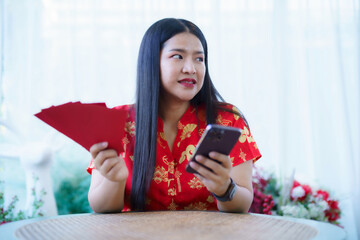 Portrait of asian woman wearing red cheongsam dress holding red envelopes and smartphone at home in the living room indoor decoration for Chinese new year festival celebrate culture of china holiday
