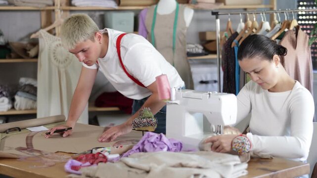 Attentive woman tailor sewing textile using sewing machine and young male assistant drawing pattern on sheet of paper using curves in fashionable sewing atelier