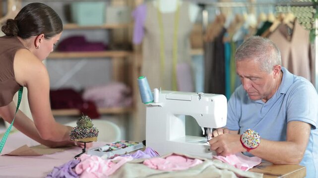  Working day in sewing workshop, textile industry. Mature man master sew clothes in sewing machine, girl pins paper pattern to fabric, attaches template to fabric, preparing to cut fabric into pieces.