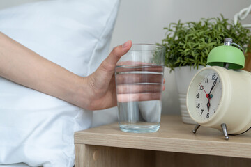 Close up of woman sitting on bed, holding a glass of water in bedroom.