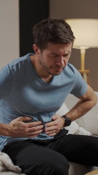 Man sitting on sofa in living room holding stomach and suffering from abdominal pain or cramps. Person suffering from health issues, digestion problems or food poisoning at home