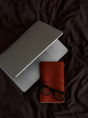 Laptop, leather cover notebook, glasses on brown bed linen