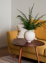 The interior of the living room - a yellow sofa, a wooden table, a ceramic vase with juniper branches. A cozy house