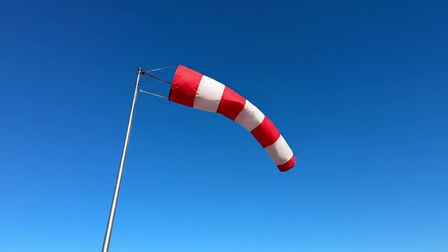 A vibrant red and white striped windsock billows gracefully against a clear, expansive blue sky on a bright, sunny day. The fabric, extended horizontally and taut by the force of the invisible wind, c