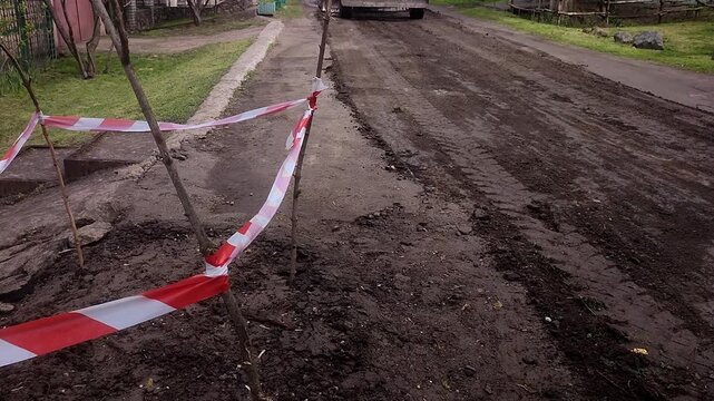 Road repair in a private residential area, on a street between single-story houses. Heavy equipment laying asphalt. A grader and asphalt paver.