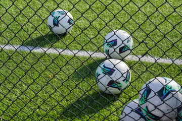 a cluster of white soccer balls resting on green artificial turf near a white field line, viewed through the diamond mesh of a black chain-link fence.