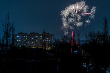 A beautiful fireworks display illuminates the night sky above a city apartment building silhouette. The vibrant sparks create a dazzling starburst pattern, capturing a festive celebration atmosphere.
