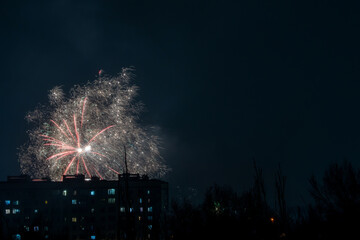 A beautiful fireworks display illuminates the night sky above a city apartment building silhouette. The vibrant sparks create a dazzling starburst pattern, capturing a festive celebration atmosphere.