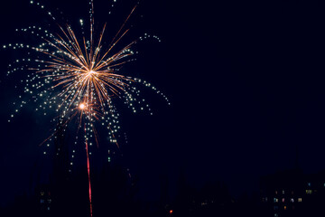 A beautiful fireworks display illuminates the night sky above a city apartment building silhouette. The vibrant sparks create a dazzling starburst pattern, capturing a festive celebration atmosphere.
