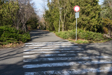 a paved park road featuring a white striped crosswalk in the foreground, flanked by green trees and bushes, with a red traffic sign standing on the right.
