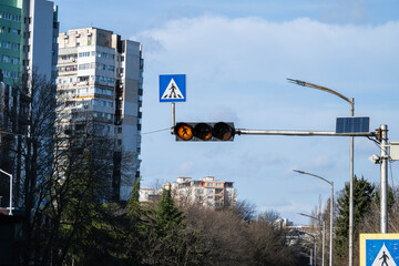 a traffic light assembly suspended over a city street, featuring a flashing yellow pedestrian warning light and a blue square pedestrian crossing sign, with tall apartment buildings in the background.