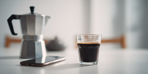 Freshly brewed espresso in a glass next to a classic moka pot and smartphone on a white table, modern morning routine and home office concept in a bright minimalist setting