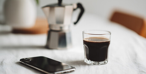 Glass of black coffee and smartphone on a white tablecloth with a blurred moka pot in the background, morning routine and lifestyle concept for remote work
