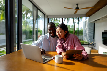 Multinational couple enjoying their morning routine, smiling and using laptop and tablet at home