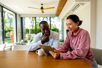 Multinational couple enjoying their morning routine, smiling and using laptop and tablet at home