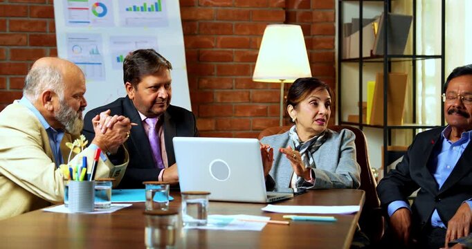 Indian businesswoman or boss briefing team as senior Indian Asian female leader addresses senior management sitting around conference table with laptop computers inside modern corporate office