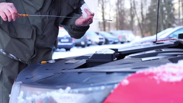 A man checks the oil level in a car engine during winter. Snow covers the ground, and parked cars are visible in the background.