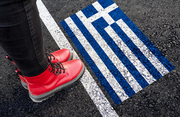 a woman with a boots standing on asphalt next to flag of Greece and border
