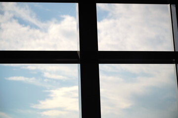 建物の窓枠越しに眺める青空と白い雲のコントラストが美しい抽象的な構図の写真です。黒いフレームが空の広がりを強調し静かな室内からの視点を表現しています。