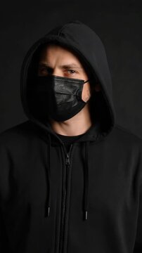 Portrait of a person wearing black hooded sweatshirt and black medical face mask against a dark background in studio setting.