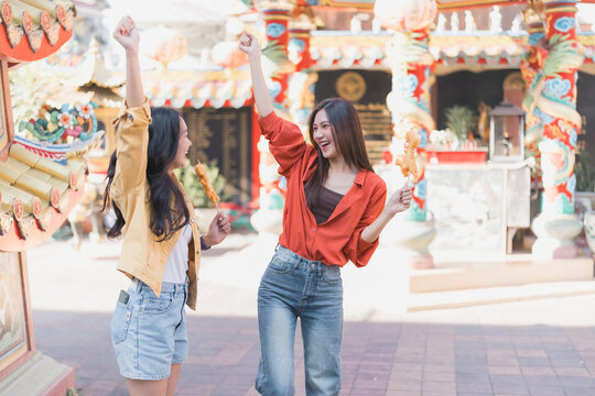 Two young Asian women joyfully raising arms and holding snacks on sticks, exploring a vibrant Chinese temple area while enjoying delicious street food together