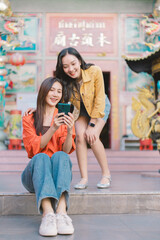Two cheerful young Asian women are sitting and standing, both looking at a smartphone screen, sharing a moment and smiling happily in front of a traditional temple building