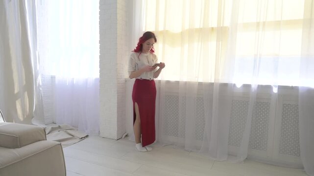 Young red-haired woman using phone by bright window. Red maxi skirt, white top. Modern, relaxed, minimalist, sunlit home interior. Digital lifestyle.