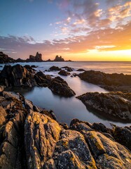 A coastal scene at sunset featuring jagged rock formations. Calm water reflects the vibrant sky with fiery hues