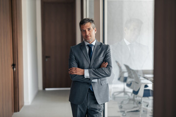 Fototapeta premium Confident Businessman In Grey Suit Stands With Arms Crossed In Office Corridor