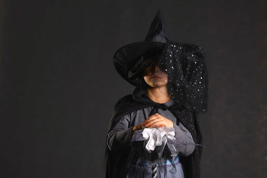 Girl in classic witch costume with pointed hat and veil standing with hands clasped in dark studio, costume for Halloween or carnival