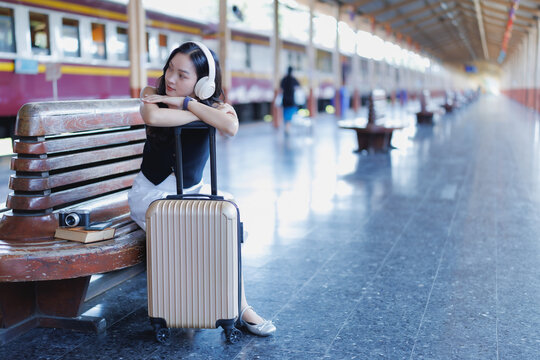Young Asian woman sitting on a bench at a train station platform, wearing headphones and leaning on her luggage while waiting for a train and listening to music