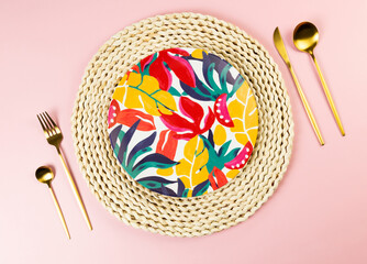 Top view of brown wicker table mat, colorful plate, gold cutlery on pink background. Straw round...