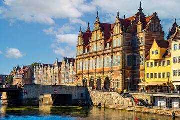 Fototapeta premium Charming historic architecture lines the waterfront of Gdansk under a clear blue sky. The river is bustling with boats, offering a vibrant scene with the city's iconic brick buildings