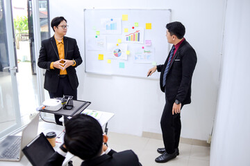 Two Businessmen In Suits Presenting Data On Whiteboard To Colleague In Modern Office Setting